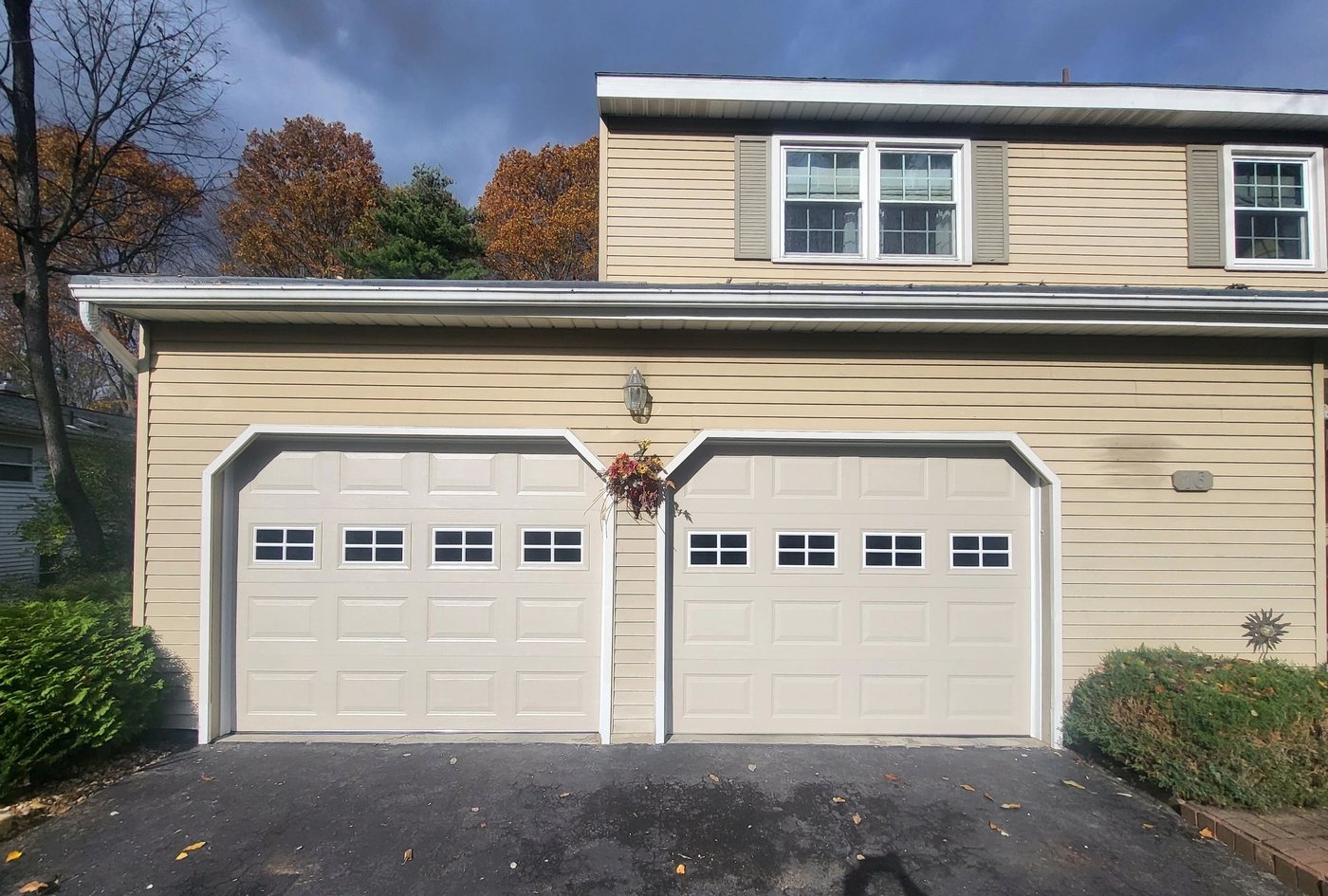A matched pair of almond doors with square window rows, set into white clipped-corner surrounds on a tan two-story, autumn trees behind.